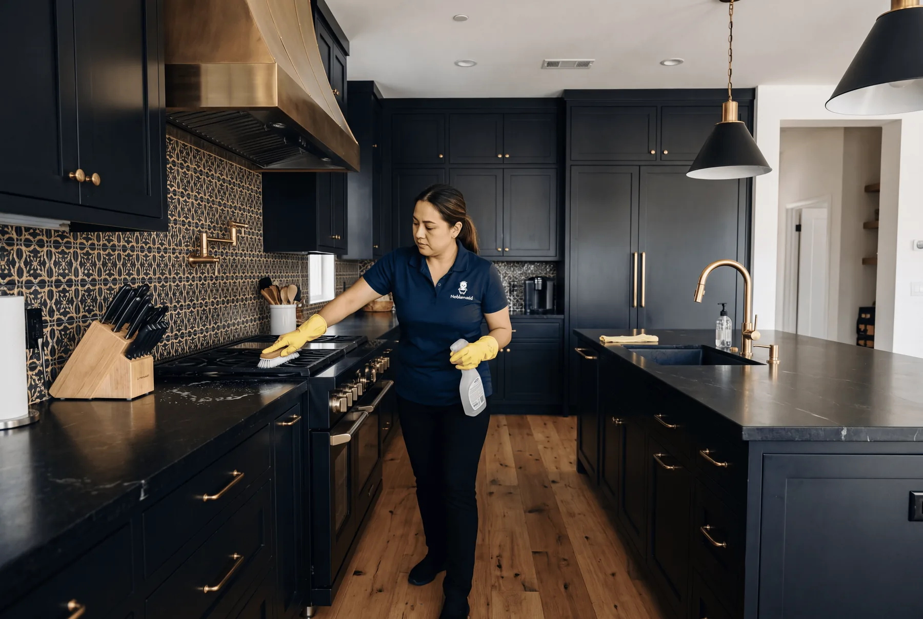 A Noblemaid cleaner scrubbing a gas range in a navy kitchen during a deep clean