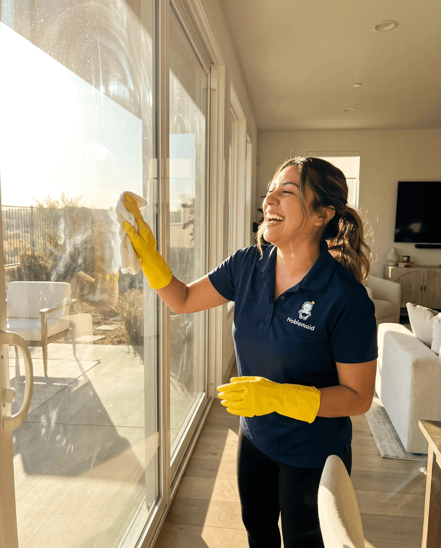 A Noblemaid cleaner in a navy polo wiping a sunlit sliding glass door